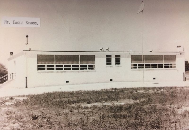 Black and white photograph of the four-room Mount Eagle Elementary School. The side of the building facing North Kings Highway is pictured. Each classroom has a large bank of windows. There is an entrance on the side of the building, with concrete steps and metal railings. An American flag is flying from a flag pole in front of the structure. 