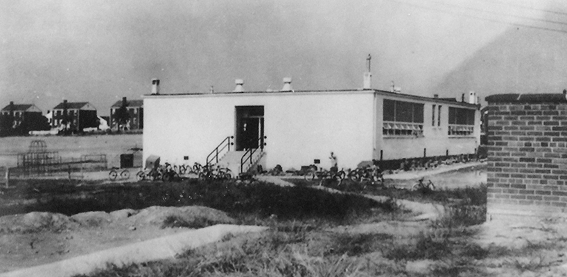Black and white photograph of the four-room Mount Eagle Elementary School. This photograph is taken from an angle where the church building stands today, and was donated by the church which used the school for their Sunday School classes. There are many, many bicycles on the ground next to the building. In the rear of the building there is some playground equipment visible. Houses can be seen in the far distance. 