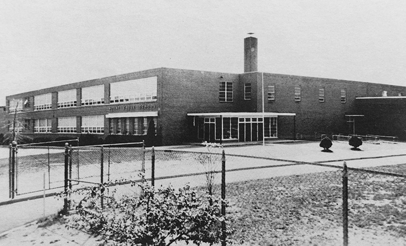 Black and white photograph of Mount Eagle Elementary School, dated 1957, from a yearbook. The photograph was taken from the sidewalk along North Kings Highway. The building looks very similar to the 1954 photograph, except the administrative wing of the building that led to the cafeteria has been enlarged to two-stories tall and the wing now extends toward the rear of the building. A chain link fence has been installed around the school grounds.  