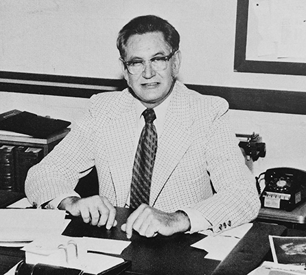 Black and white yearbook photograph of Principal Tarbox. He is seated at his desk with papers in front of him. A rotary dial telephone is visible in the background.  