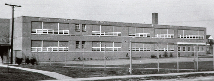 This is an animated gif image showing the front of Mount Eagle Elementary School. The background photograph is a black and white image of the building taken in 1958 from the south end of the building showing only the front two-story classroom wing. It was taken from relatively the same angle as the earlier photograph of the 1949 building, so the 1949 picture has been superimposed over top of the 1958 image. The 1949 school fades in and out, showing where it was located in relation to the enlarged structure. The windows and their placement are identical in each image. 